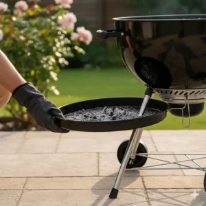A person wearing a protective glove removing the ash catcher tray from beneath a kettle BBQ for easy cleaning.