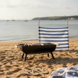 A small portable charcoal BBQ set up on a sandy beach with a windbreak in the background.