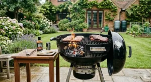 A close-up of a wooden side table featuring British BBQ essentials: a jar of Tracklements mustard, a dry rub shaker, a basting brush, and a can of Meantime London Pale Ale.
