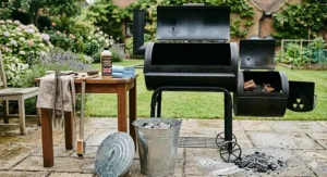 A cleaning setup featuring a galvanised ash bucket, a wire brush, and a bottle of specialist grate cleaner next to the open smoker, illustrating essential maintenance for a charcoal barbecue.