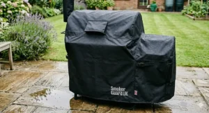 The offset smoker fully protected by a heavy-duty, waterproof black cover with a Smoker Guard UK logo, standing on a wet stone patio to demonstrate weather resistance for the British climate.