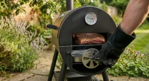 A hand in a protective heat-resistant glove adjusting the circular air intake damper on an offset barbecue to regulate cooking temperature.