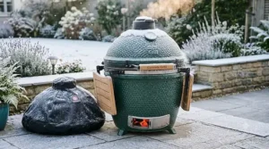 A robust kamado BBQ grill with its protective cover, dusted with light snow in a British garden, highlighting year-round build quality.