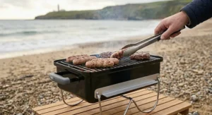 A close-up of sausages and burgers being turned with metal tongs on a small charcoal grill, with a scenic coastal view in the background.