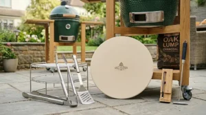 An array of essential kamado BBQ accessories, including stainless steel tongs, a pizza stone, and a temperature probe, displayed on a patio.