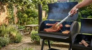 Using stainless steel tongs to place a piece of seasoned brisket onto the porcelain-coated cooking grates of an offset smoker.