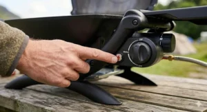 A close-up photograph of a finger pressing the red piezo ignition button on the control panel of a black portable gas BBQ, which is resting on a wooden picnic table.