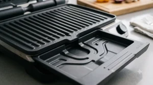 Detailed product shot showing the dark, non-stick ribbed grilling surface and the open, sculpted grease collection tray of a modern electric barbecue, highlighting easy cleaning features.