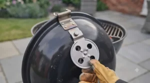 A hand in a glove adjusting the metal air vent on the lid of a charcoal kettle BBQ to manage cooking temperature.