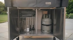 A view of the barbecue trolley with its double cabinet doors open, revealing a neatly organised interior complete with a spare gas cylinder, cleaning brushes, and stored tools, highlighting practical storage solutions.