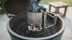 A metal chimney starter filled with glowing lumpwood charcoal resting on the grate of a black kettle barbecue.
