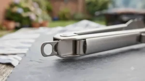 An extreme close-up photograph focusing sharply on the hinge and pull-tab locking mechanism of the stainless steel BBQ tongs, highlighting the brushed metal texture and precise engineering.
