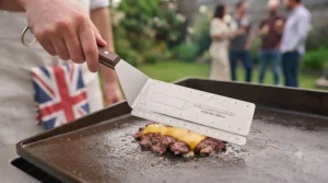 Chef using a wide spatula for burgers to press a mince patty flat onto a hot cast-iron skillet